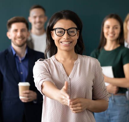 Mixed race woman sales manager stretch out hand introduces herself greeting shake hands client smiling look at camera pose indoors with diverse teammates. HR, job interview, business etiquette concept